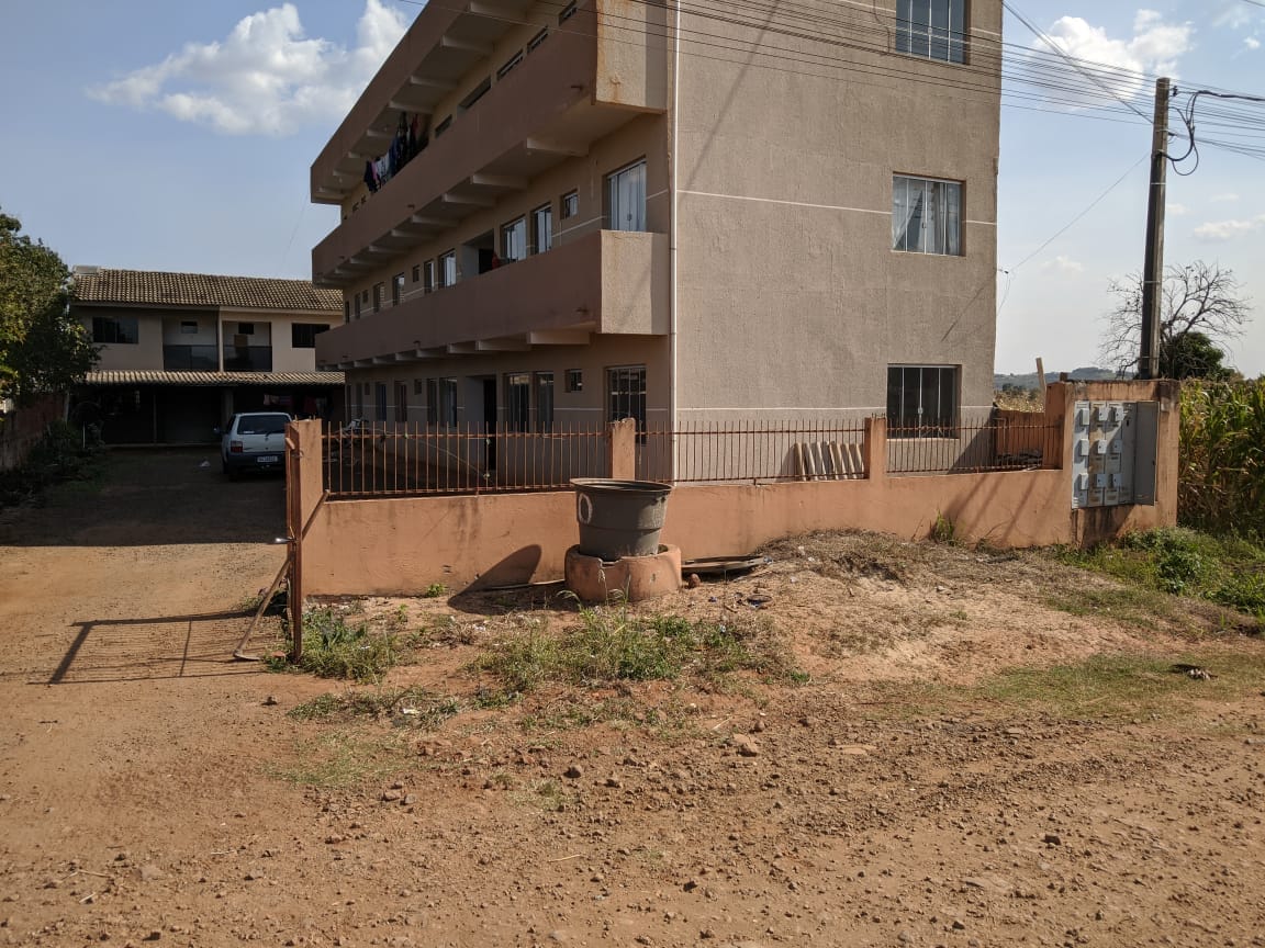The exterior view shows a three-story building with a simple, functional design. A gravel driveway leads to the entrance, bordered by a low fence. A few vehicles are parked nearby, and the surrounding area features sparse vegetation and open space.