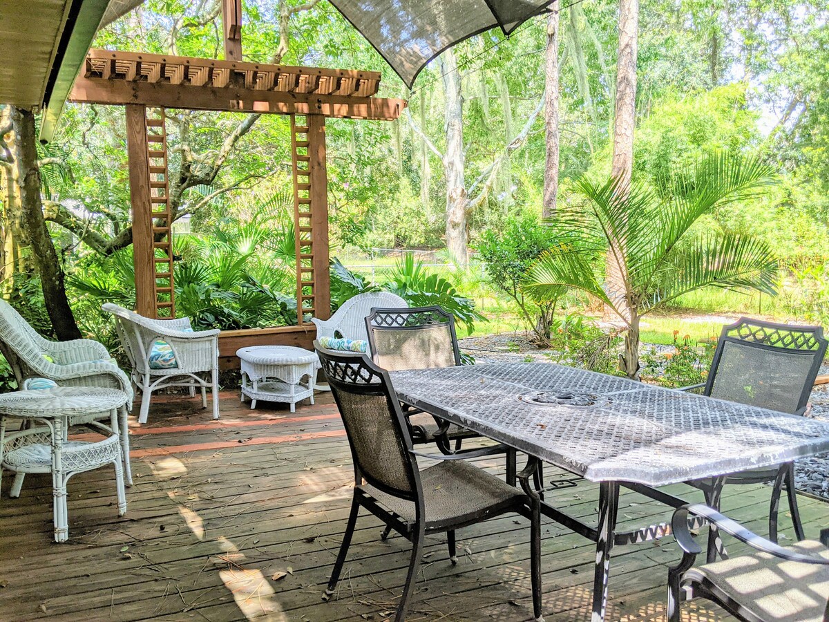 An outdoor seating area features a round table with a center fire pit and several surrounding chairs. Wicker furniture offers additional seating under a shaded pergola. Lush greenery, including palm trees and various plants, frames the space, creating a tranquil setting for relaxation.