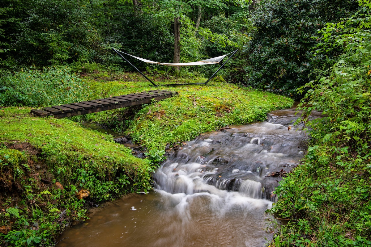 A tranquil scene of a hammock set between trees, adjacent to a gently flowing stream. The lush green surroundings feature manicured grass and a small wooden bridge crossing over the water, inviting moments of relaxation and connection with nature.