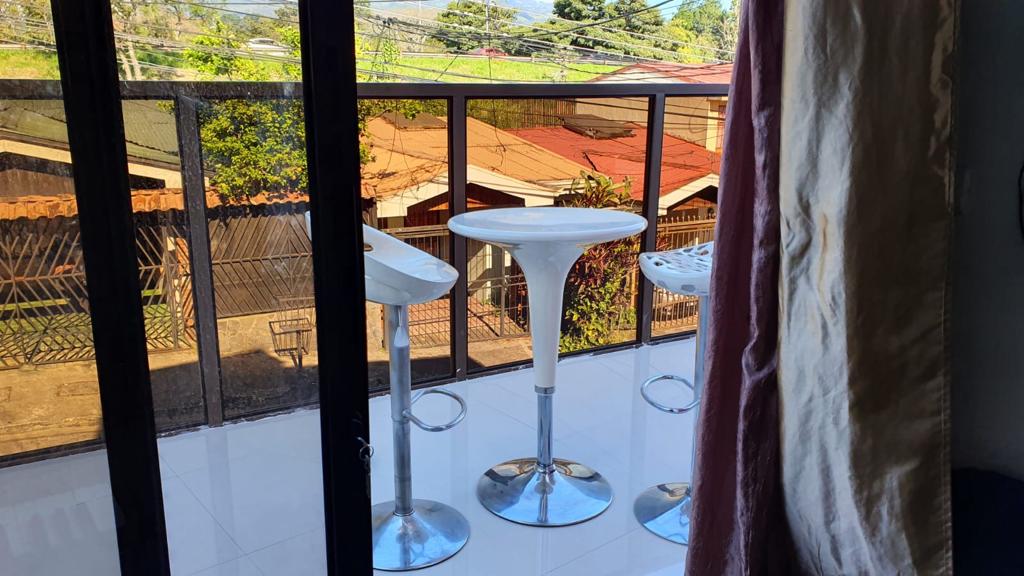 A balcony is shown with three modern bar stools positioned around a small table. Floor-to-ceiling glass doors provide a clear view of the surrounding area, enhancing the connection between indoor and outdoor spaces while allowing natural light to fill the room.