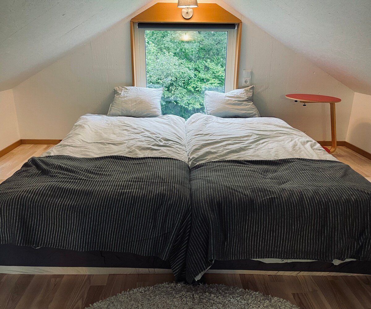 A spacious loft sleeping area features a pair of beds with soft bedding, positioned near a large window that offers a view of greenery outside. A small side table is located next to the beds, adding functionality to the cozy space.
