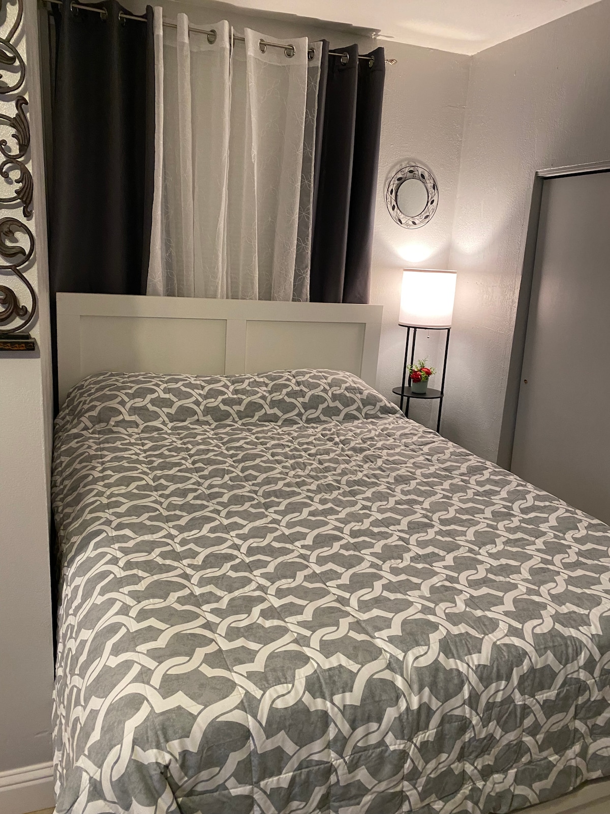 The sleeping area features a queen size bed with a gray patterned comforter. Light is provided by a small lamp next to the bed. Dark curtains frame a nearby window, and a mirror is mounted on the wall opposite the bed.