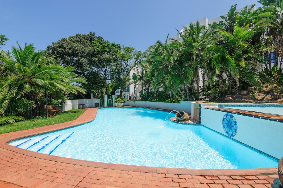 A spacious pool area is surrounded by tropical landscaping, featuring lush palm trees and manicured greenery. The pool has a unique curved shape, with clear blue water reflecting the sunlight. A relaxing atmosphere is created by the inviting poolside colors and natural elements.