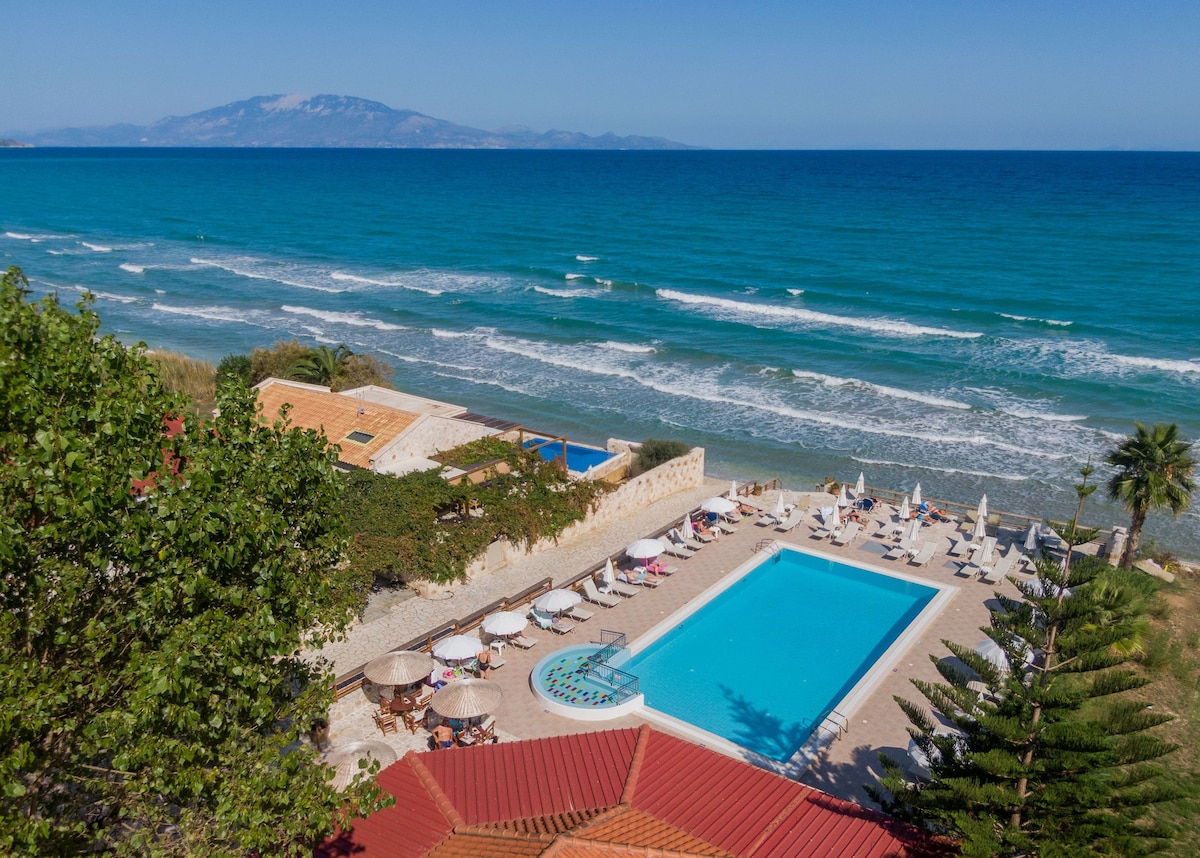 An expansive swimming pool is situated near the beach, overlooking the sparkling sea. Sunbeds and parasols are evenly arranged around the pool area. Lush greenery enhances the surroundings, while distant mountains create a scenic backdrop against the clear sky.
