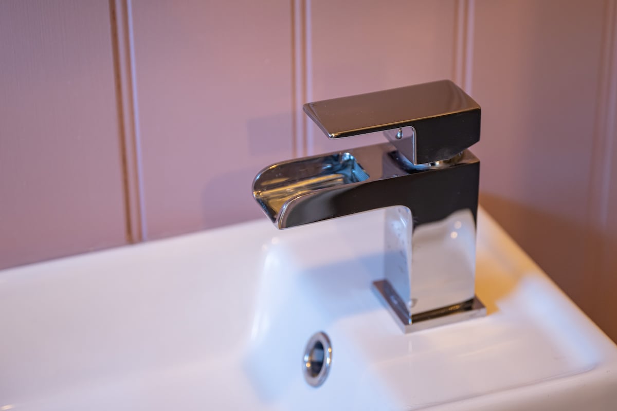 A modern faucet is positioned above a white sink, featuring a sleek chrome finish that reflects the surrounding light. The faucet's spout is elegantly designed, and the sink is set against a backdrop of soft, muted pink paneling.