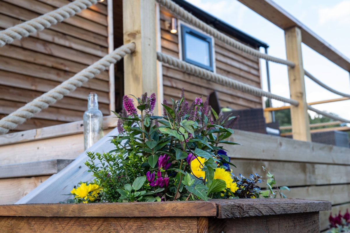 A wooden planter overflowing with a variety of colorful flowers is positioned in the foreground. The rustic exterior of the shepherd's hut is visible in the background, featuring a window and a railing made of rope. Natural wood tones create a harmonious connection with the environment.