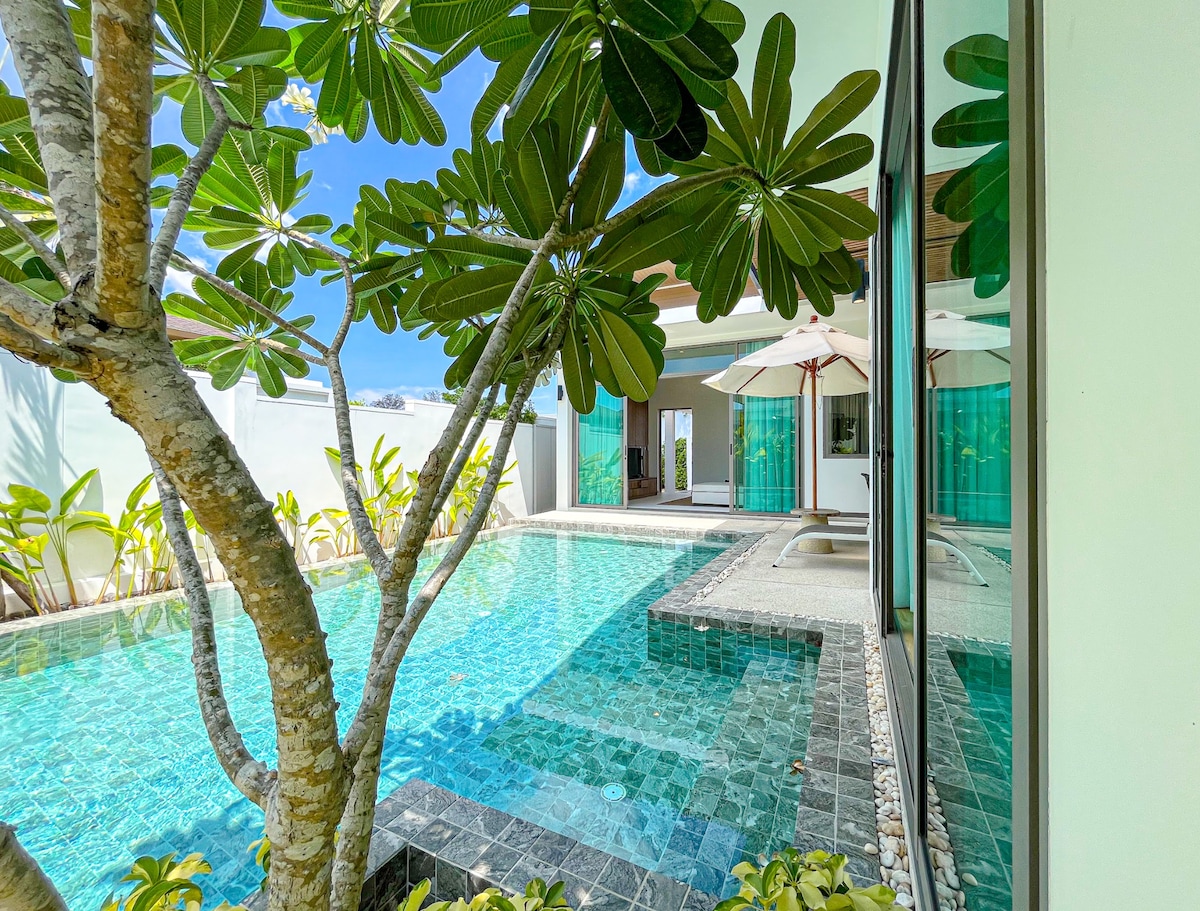 A private pool area is framed by lush greenery and tropical plants. The clear water reflects the bright sky, while a shaded lounge space is visible in the background. Sliding glass doors provide a view into the villa's interior, enhancing the seamless connection between indoor and outdoor living.