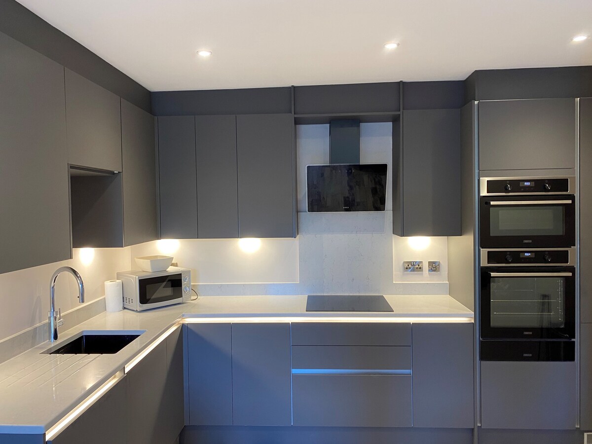 A modern kitchen features sleek gray cabinetry and a white countertop. Built-in appliances, including a microwave and ovens, are installed beneath overhead cabinets. Soft lighting illuminates the space, highlighting the minimalist design and a sink integrated into the counter.