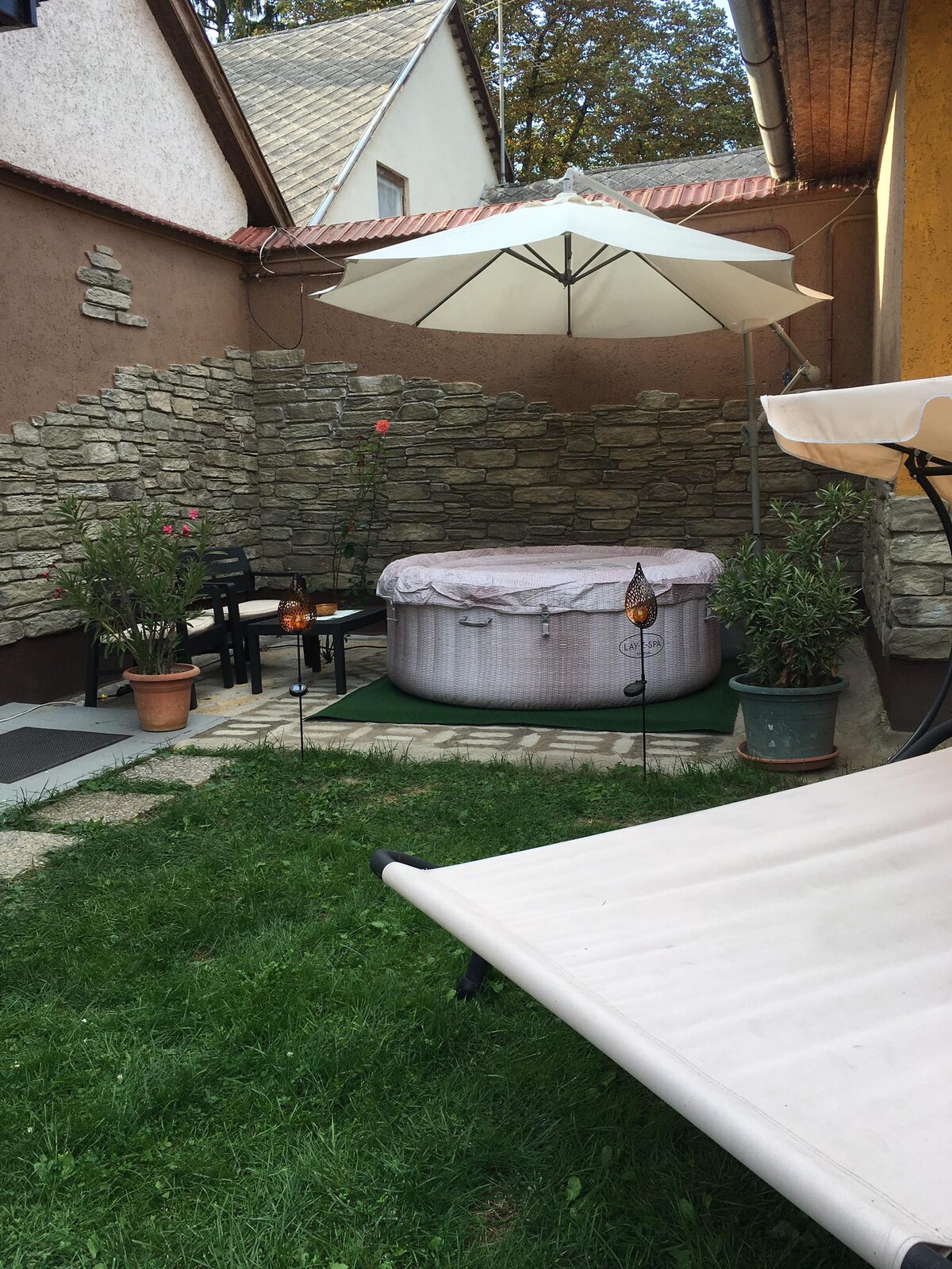 A private outdoor space is featured with a jacuzzi surrounded by stone walls. Potted plants and a pair of chairs are visible along with a large umbrella providing shade. A sun lounger sits on the grass, inviting relaxation in this tranquil setting.