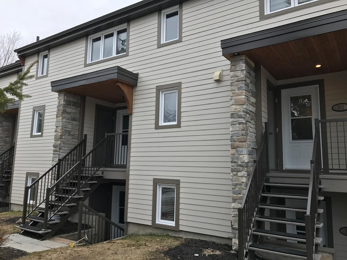 The exterior of a two-story townhouse is displayed, featuring a light-colored siding complemented by stone accents. Staircase entrances lead to individual units, with large windows allowing natural light to enter. A grassy area is visible in front, contributing to the home's inviting appearance.