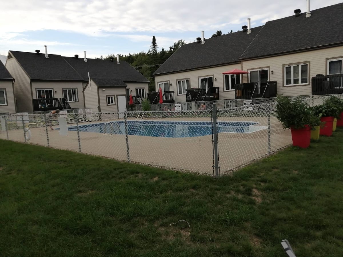 The outdoor pool area is surrounded by a chain-link fence, featuring several lounge chairs and umbrellas for shade. Red planters with greenery add vibrancy to the concrete space. The adjacent townhouse units offer access to the pool, creating a relaxed environment.