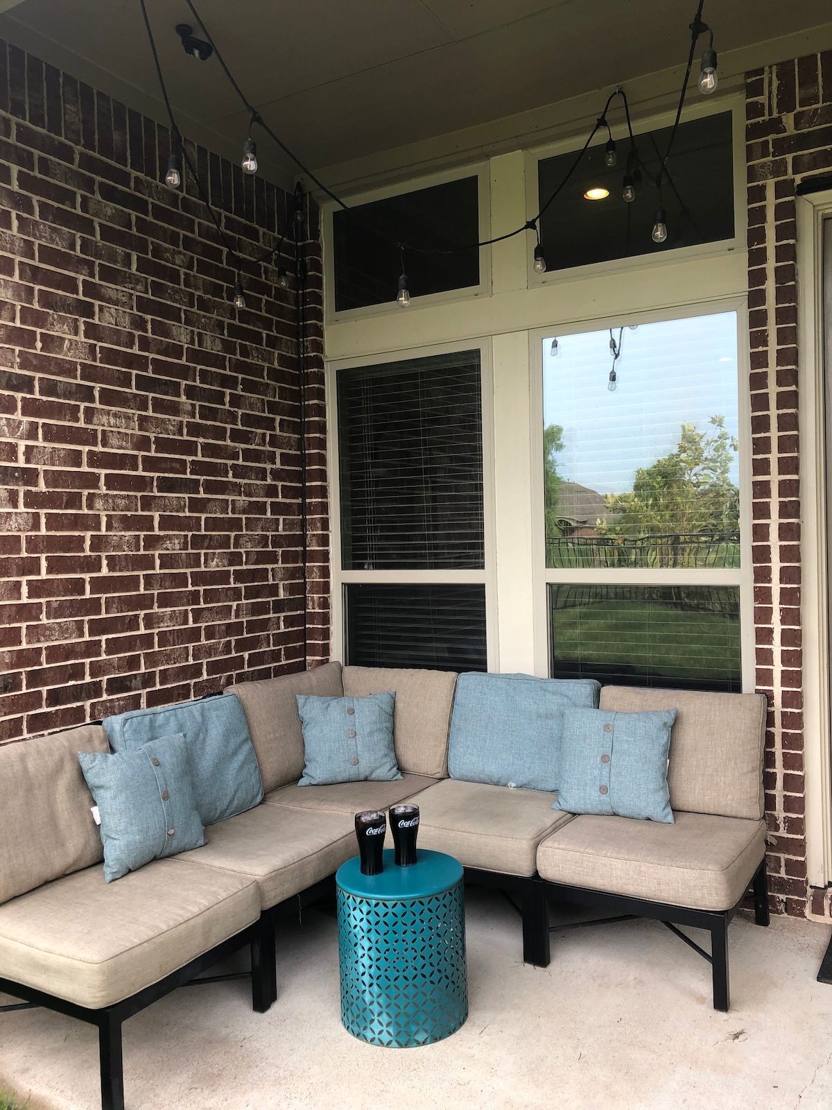 A cozy outdoor seating area is created with a sectional sofa adorned with blue cushions. A small turquoise accent table sits in the center, while soft string lights hang above, illuminating the space. Large windows provide a view of the lush exterior.