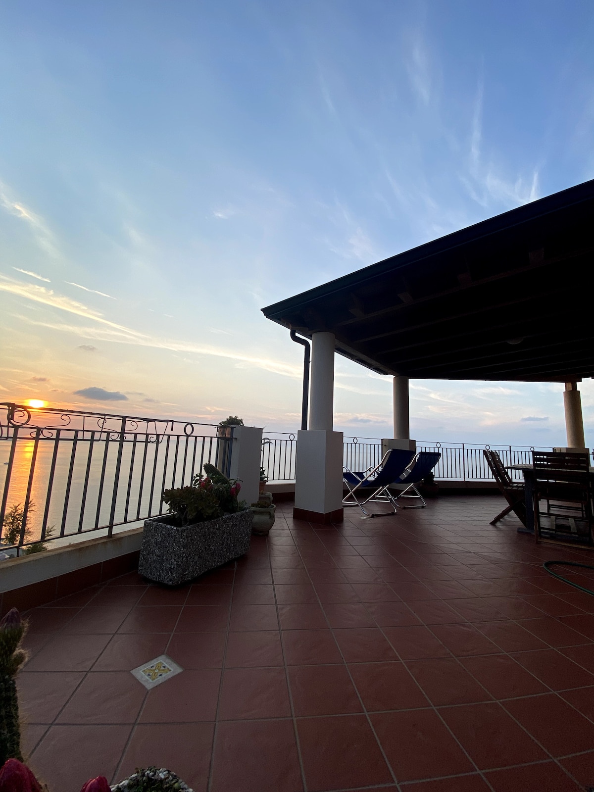 A spacious terrace is shown, featuring a partial roof that offers shade. Two reclining chairs are positioned for relaxation, with a dining area visible in the background. A planter with greenery adds a natural element, while a scenic sunset over the horizon provides a calming backdrop.