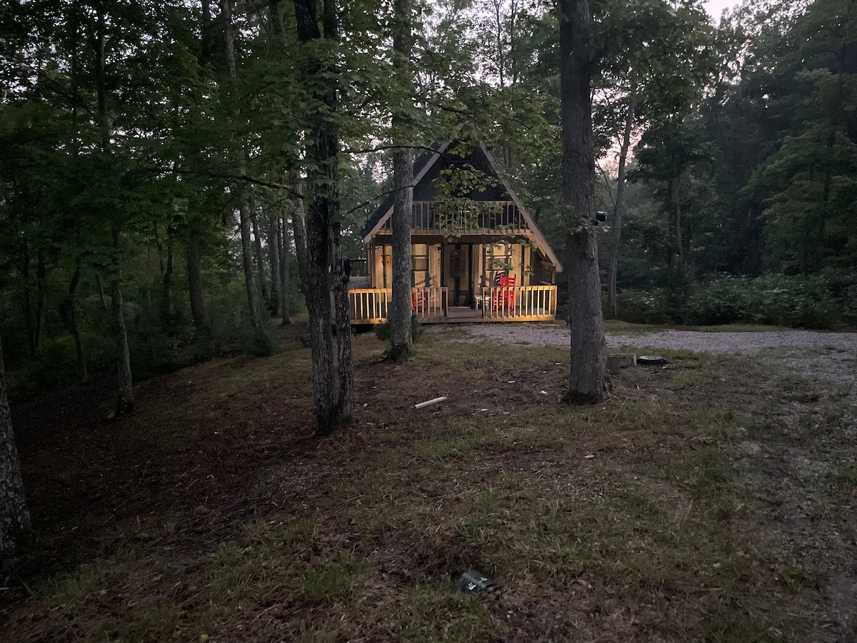 An A-frame cabin is nestled among trees, featuring a welcoming porch with wooden railings and several chairs positioned in front. The cabin's exterior is partially illuminated, with warm light visible through large windows. The surrounding area is grassy and natural, creating a serene outdoor setting.
