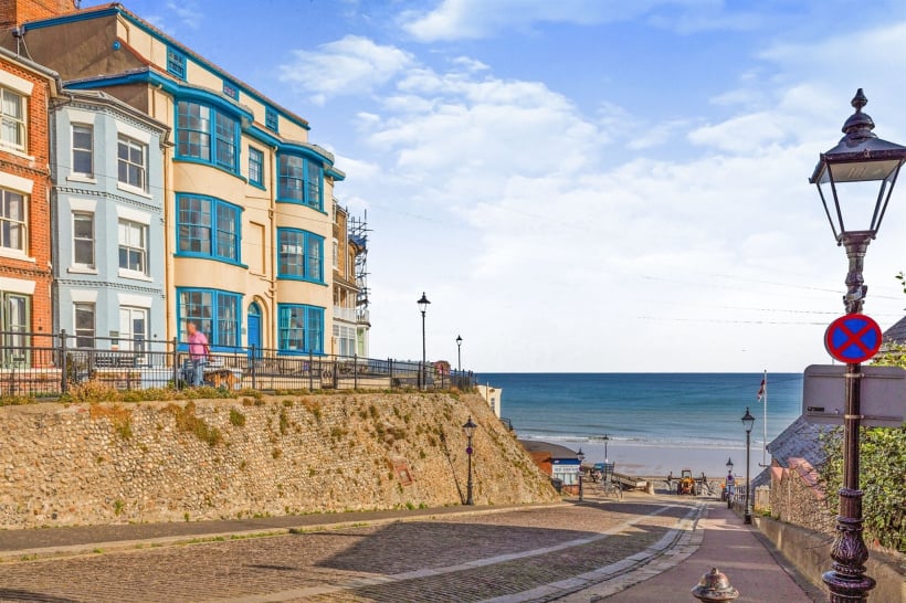 A scenic view is presented, showcasing a sloping pathway leading to the beach. The coastline is visible in the background, with the sea meeting the sky. A row of colourful buildings lines the walkway, along with street lamps, enhancing the vibrant atmosphere.