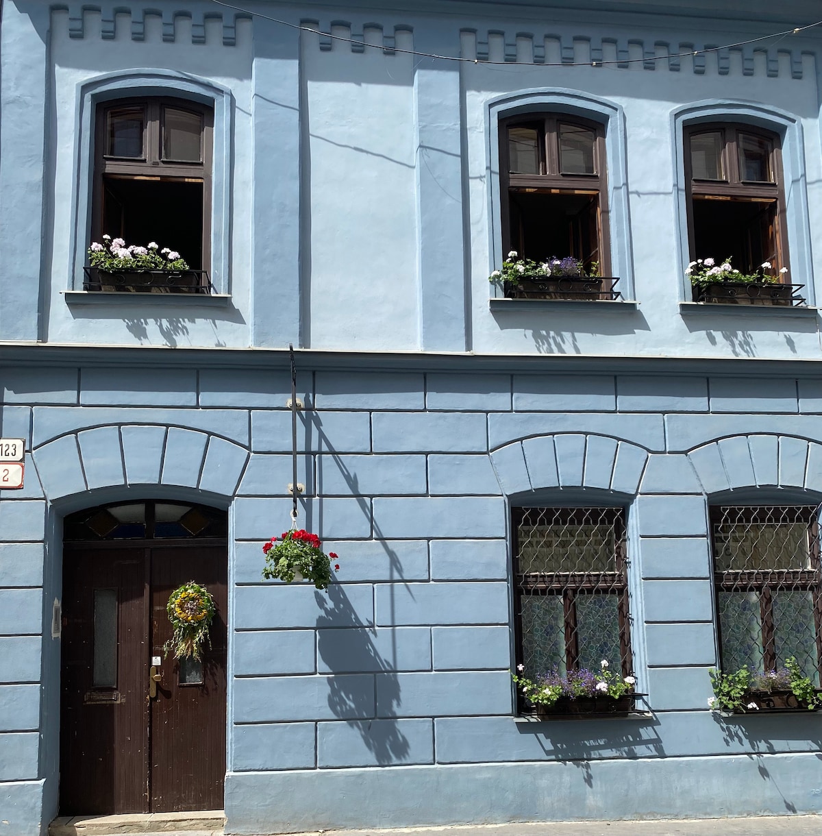 A charming building exterior is painted in soft blue, with dark wooden window frames highlighting flower-filled window boxes. A decorative wreath adorns the front door, which is flanked by two arched windows. Natural light enhances the inviting atmosphere of this historic structure.