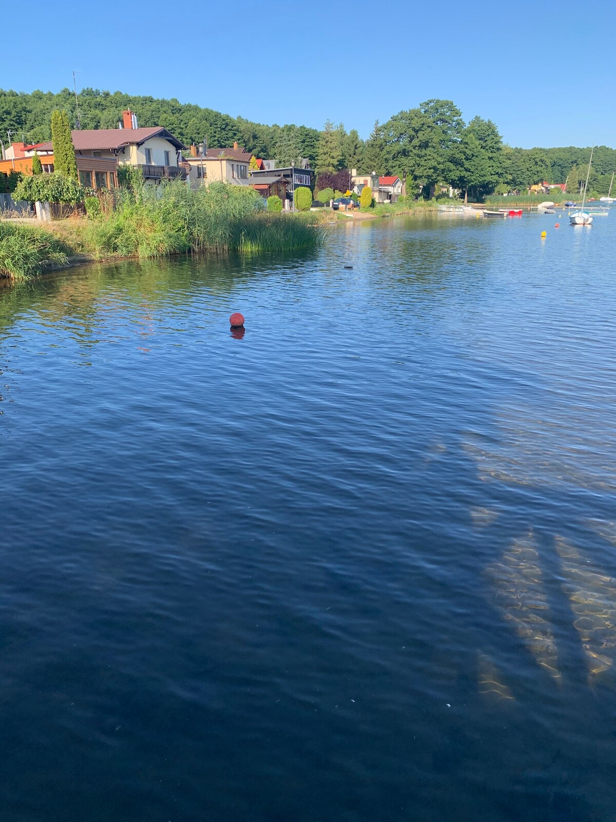 Calm water reflects the clear blue sky, with a red buoy anchored in the foreground. Lush greenery borders the shoreline, and a few boats are visible in the distance, adding to the serene lakeside atmosphere. Houses are set back along the water, contributing to the peaceful setting.