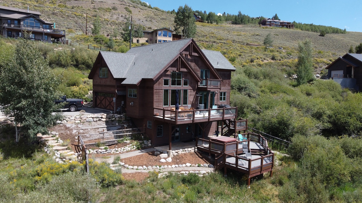 An exterior view of a custom wooden home featuring multiple decks and balconies. Surrounded by greenery, the property offers mountain views and a spacious outdoor area with landscaped stones. The structure showcases multi-pane windows and a gabled roof, embodying a rustic charm.