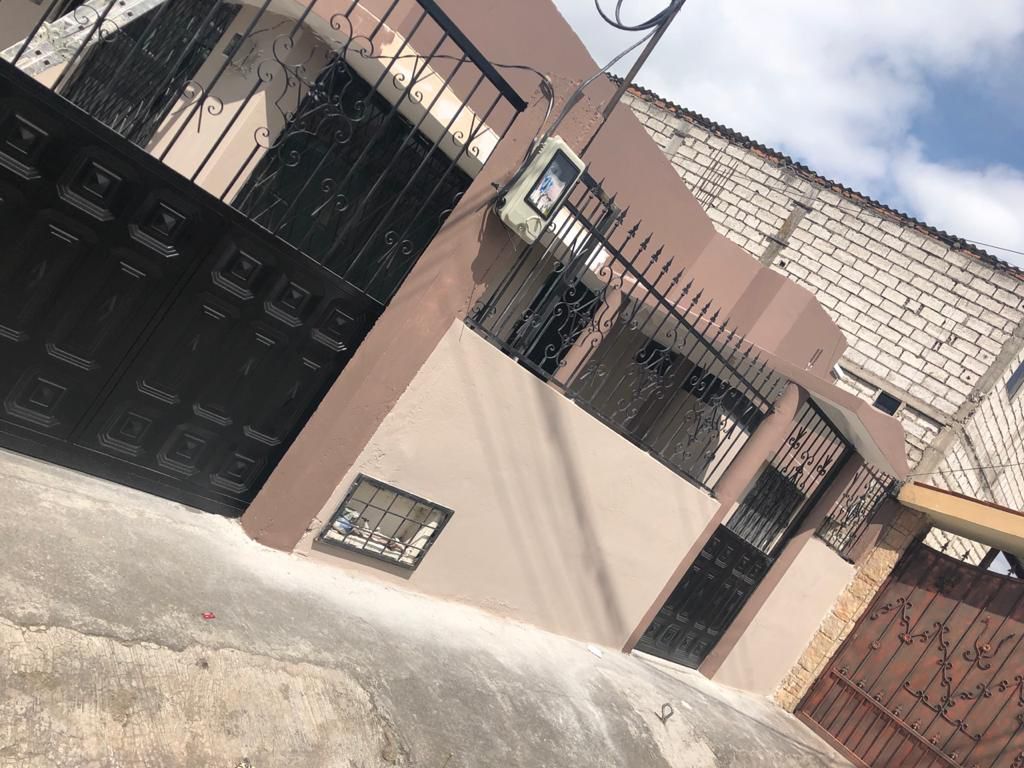 A welcoming exterior view of the property is shown, featuring a tan-colored wall with decorative wrought iron gates. The main entrance is highlighted by a solid black door, flanked by a metal fence that offers privacy. A clear sky is visible above.