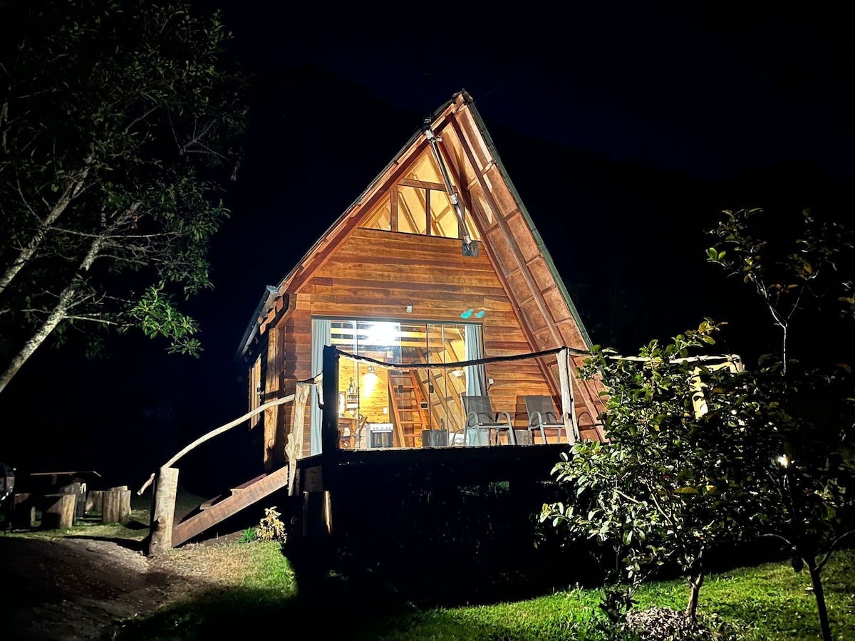 The wooden chalet is illuminated at night, showcasing its A-frame structure. A deck leads to large glass doors that reveal a warm interior. Surrounding trees and a well-maintained garden enhance the natural setting, creating a serene atmosphere.