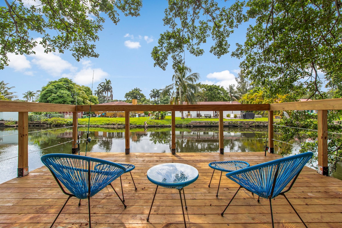 A calm outdoor deck is shown, featuring a wooden floor and four blue chairs arranged around a small table. The deck overlooks a serene canal surrounded by lush greenery and palm trees, creating a tranquil atmosphere.