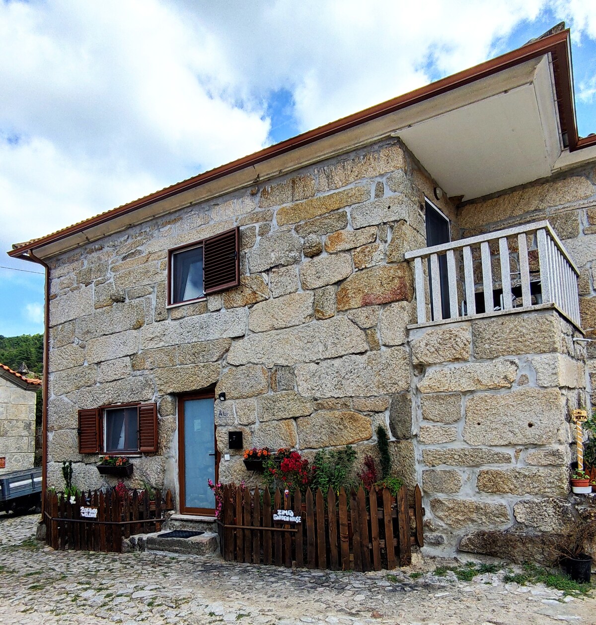 An inviting rustic house is constructed of weathered stone, featuring a light blue door and wooden shutters. A small wooden fence adorned with colorful flowers surrounds the entrance, while a quaint balcony is visible on the second floor under a partly cloudy sky.