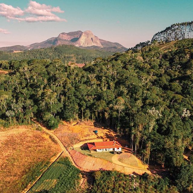 The property is positioned within a natural landscape, surrounded by lush greenery. A large, red-roofed house is centrally located, with expansive open areas and a backdrop of tall mountains under a clear blue sky.