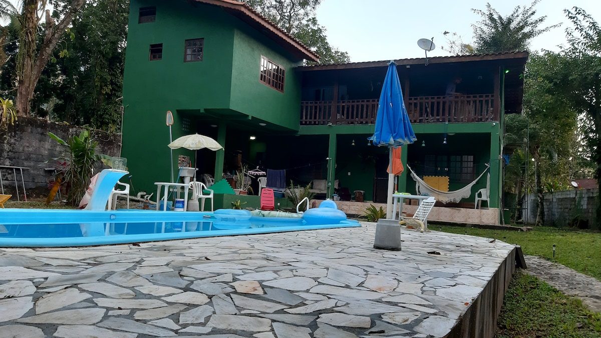 A two-story green building is situated behind a stone patio, featuring a refreshing pool in the foreground. Multiple lounge chairs and an umbrella are strategically placed around the pool area. The balcony is adorned with a hammock, creating an inviting outdoor space for relaxation.