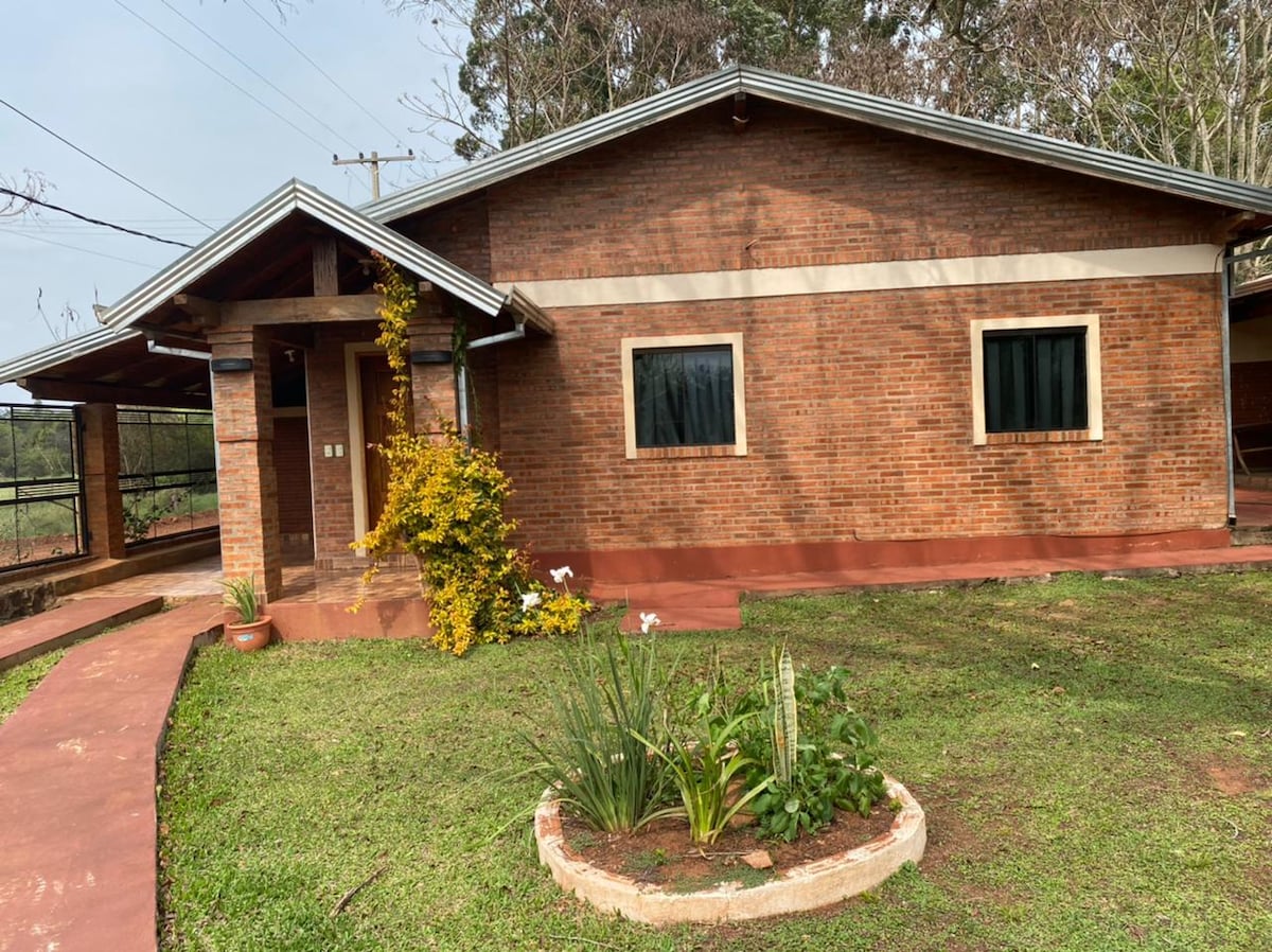 A brick cabin is surrounded by lush greenery, featuring a well-maintained garden with native plants. Two windows provide natural light, while a walkway leads to the entrance. The structure has a modern design with a roof overhang, bordered by a tranquil outdoor space.