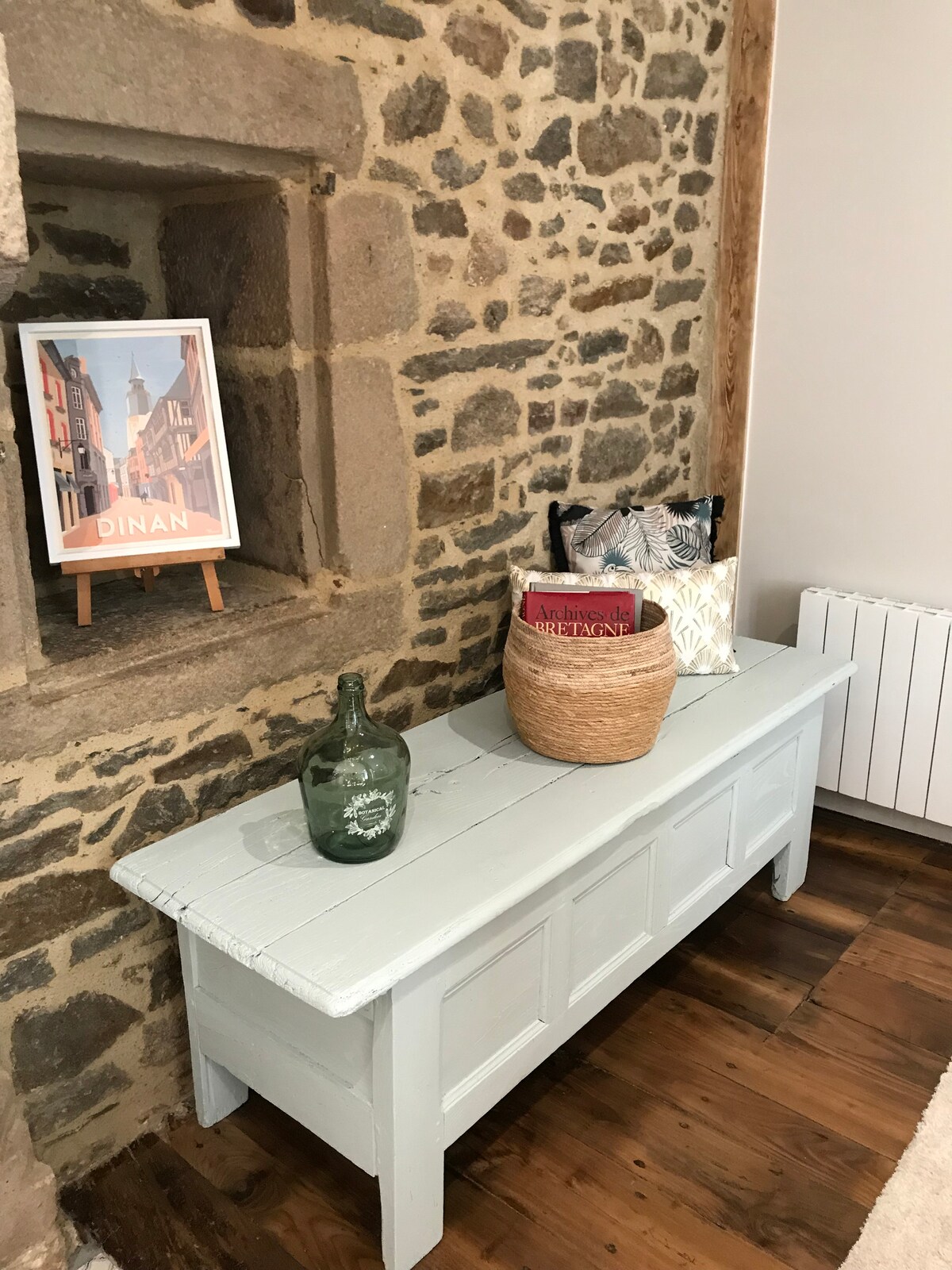 A light blue bench is positioned against a rustic stone wall, complemented by a decorative poster of Dinan. A green glass bottle and a woven basket sit atop the bench, alongside a cushion featuring monochromatic patterns. Wooden flooring adds a warm touch to the space.