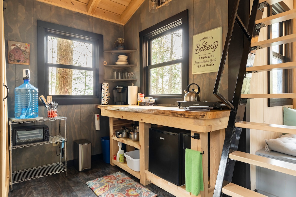 A compact kitchenette features wooden countertops, a mini fridge, and a hotplate. Shelves hold kitchenware and supplies, while a large water jug is prominently displayed. Natural light enters through windows, and a staircase leads to the upper sleeping area.