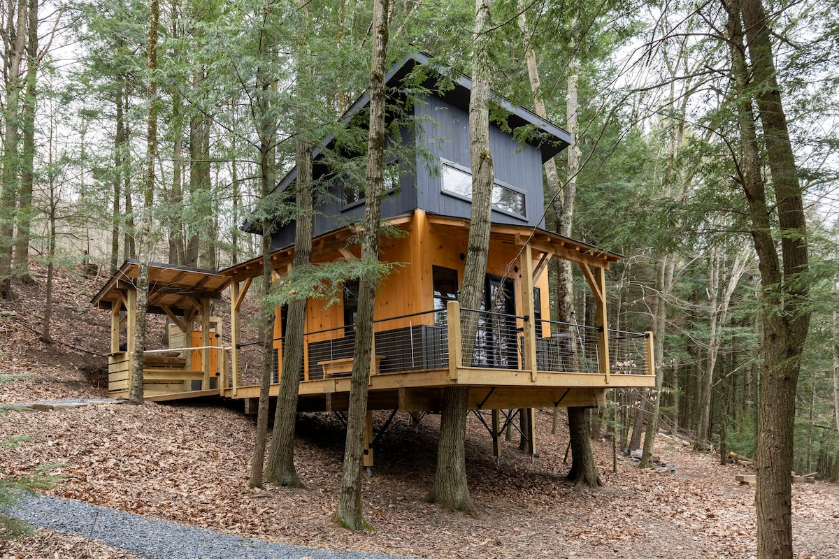 A modern treehouse is elevated on wooden stilts, surrounded by trees. A spacious outdoor deck, supported by slender posts, extends from the structure. Leafy forest floor is visible below, showcasing a natural setting with a path leading to the entrance.