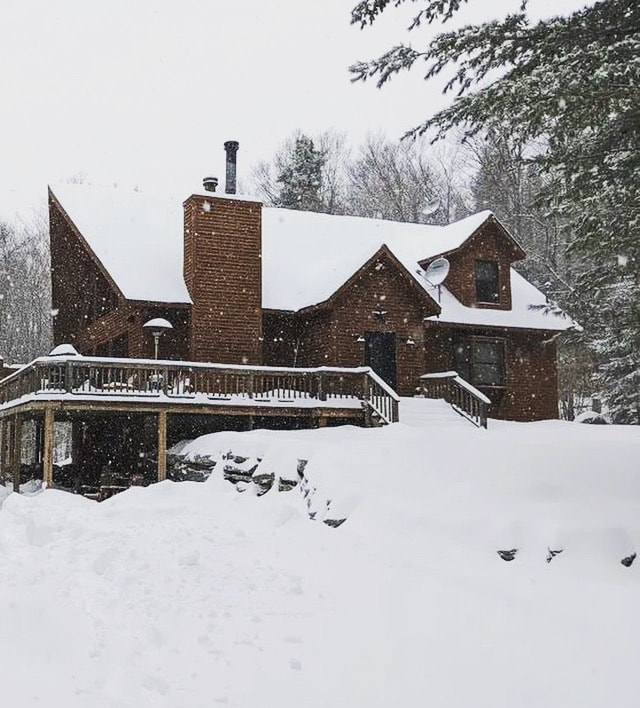 A rustic cabin is surrounded by a blanket of snow, showcasing a sloped roof with a prominent chimney. The expansive deck with railing is visible, demonstrating ample space for relaxation. Snow-covered trees frame the property, contributing to the serene winter atmosphere.