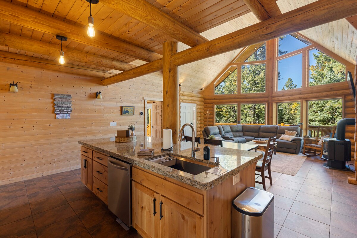 The spacious kitchen showcases wooden cabinetry and a central island with a sink and modern appliances. Natural light floods the area through large windows, highlighting the adjoining living space with a comfortable couch and views of the surrounding trees.
