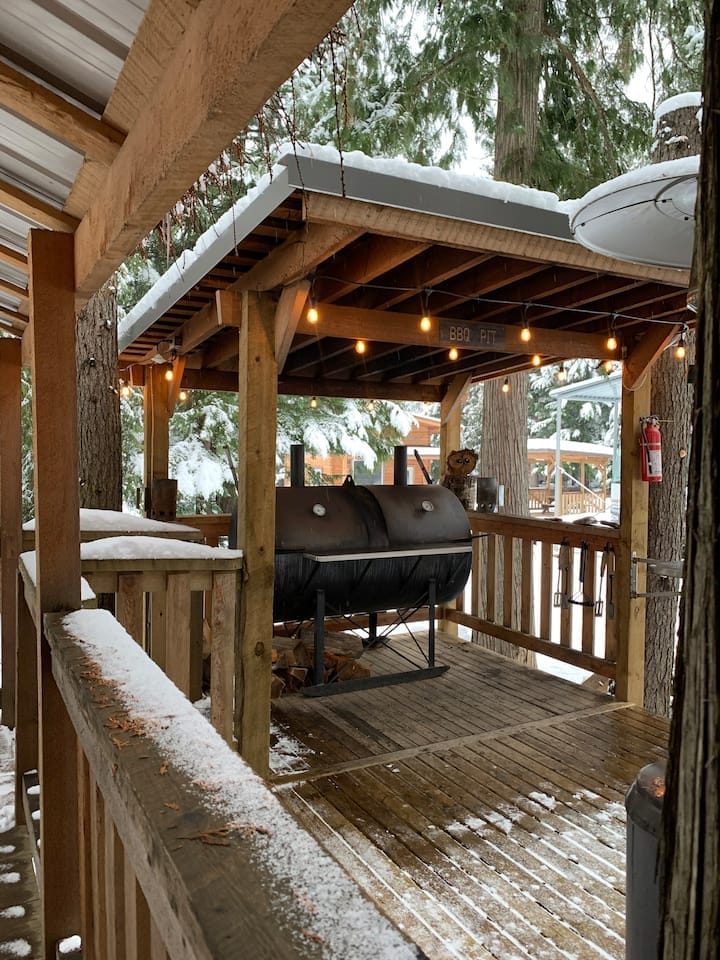 A wooden deck area is covered with light snow, featuring a custom-built BBQ/smoker positioned under a roof. String lights are hung above, creating a cozy atmosphere, with surrounding trees providing natural shade.