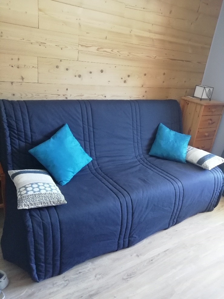 A comfortable click-clack sofa is showcased, draped in a navy cover. Two turquoise decorative pillows are positioned at either end. Wooden paneling creates a warm backdrop, and a wooden dresser is visible to the right, adding functional storage to the space.