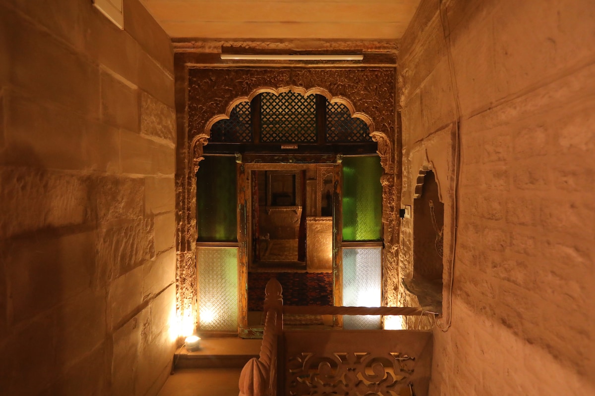 A narrow corridor is framed by intricately carved arches and warm-hued stone walls. Soft lighting highlights decorative elements, while colored glass panels create a subtle shimmer. The corridor leads to a spacious area with traditional furnishings visible in the background.