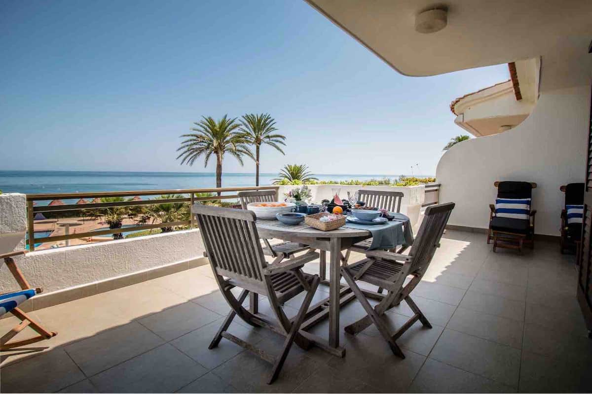 A spacious terrace features a round dining table surrounded by wooden chairs, set against a backdrop of palm trees and the ocean. The area is designed for outdoor enjoyment, with a vibrant seaside view extending to the horizon.