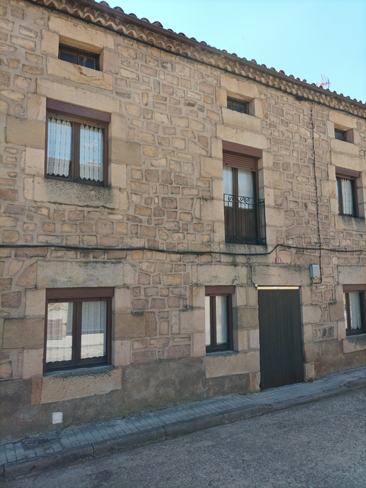 A stone-built exterior features a rugged texture, showcasing multiple windows with white curtains. A central door leads into the property, flanked by several small windows above. The structure displays a simple architectural design, contributing to a peaceful ambiance.