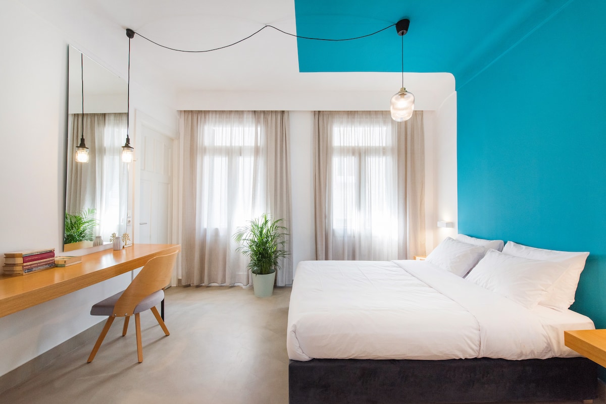 A spacious sleeping area is highlighted by a vibrant blue accent wall. Natural light filters through large windows, illuminating the minimalist furnishings, including a stylish desk and a comfortable bed. A potted plant adds a touch of greenery to the neutral color palette.
