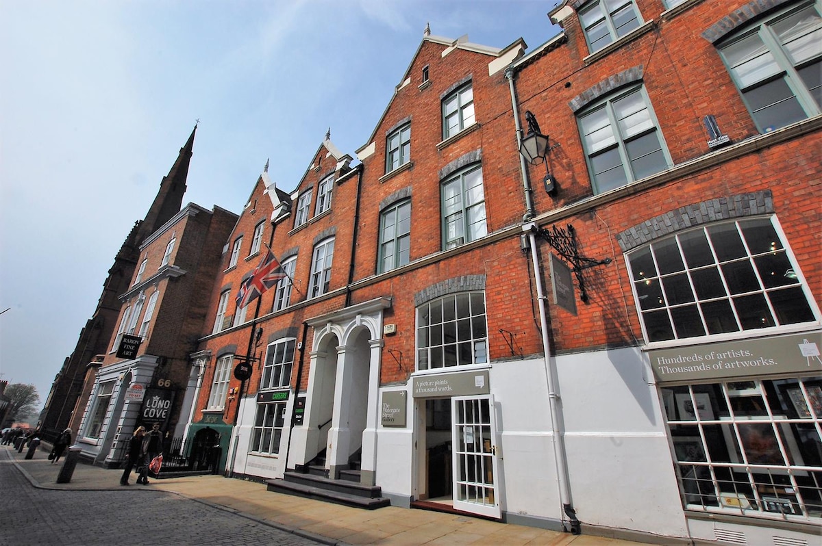 The exterior of a historic Grade 2 Listed Building showcases its red brick facade with elegant architectural details. Tall windows adorned with decorative trim are visible. The entrance features double doors framed by white stone, inviting guests into the vibrant atmosphere of Chester City Centre.