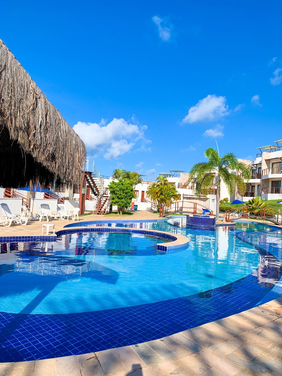 A large swimming pool, featuring distinct areas for both relaxation and play, is surrounded by lush greenery and palm trees. Sun loungers are positioned around the pool, providing ample space for guests to unwind. A shaded seating area adds comfort to the outdoor environment.