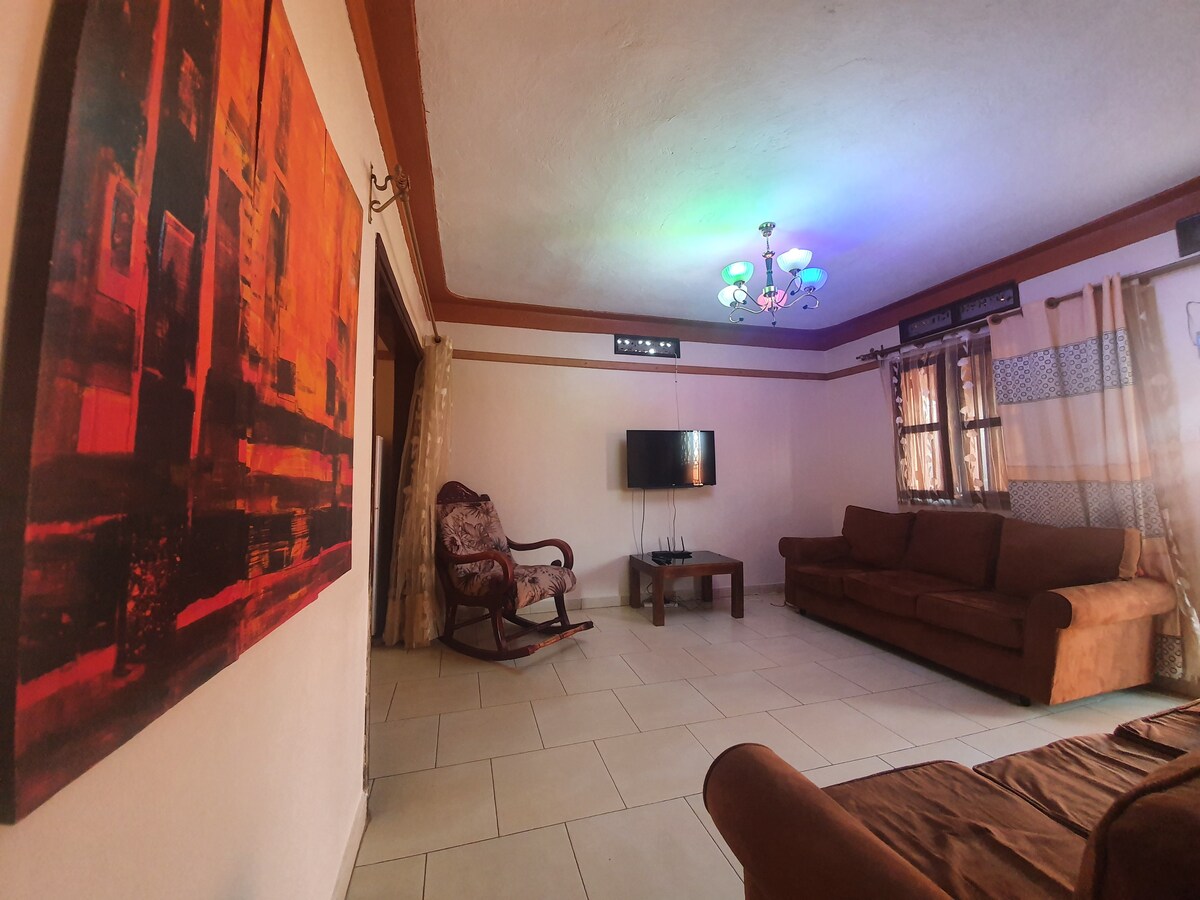 A spacious living area features a combination of soft brown furniture and natural light filtering through window treatments. A vibrant wall painting adds a pop of color, while a modern chandelier casts multicolored light throughout the room. A television is positioned on a minimalistic stand.