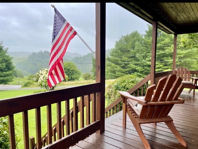 A porch view showcases an American flag gently waving in the breeze. Two wooden Adirondack chairs are positioned on the veranda, overlooking a lush, green landscape with rolling hills in the distance, framed by trees and a cloudy sky.