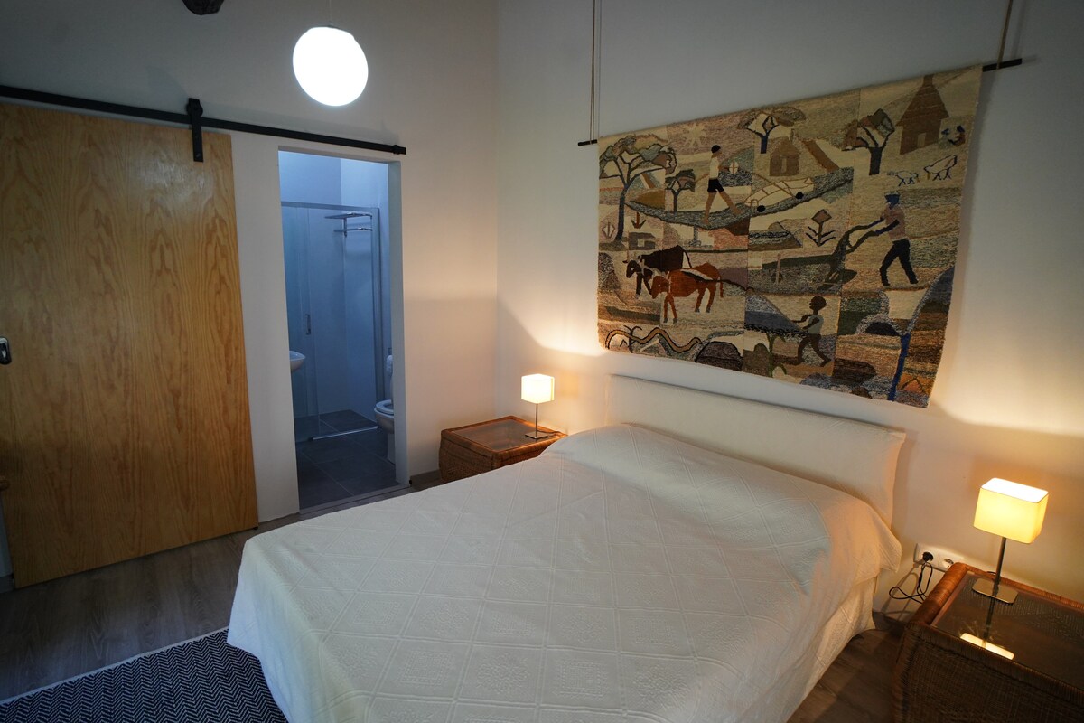 The room features a spacious bed covered with a light-colored quilt. A woven tapestry hangs above the bed, depicting a rural scene. Bedside lamps on woven nightstands provide soft lighting, while a bathroom door is visible in the background.