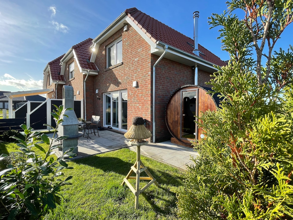 The exterior view showcases a modern brick building with a red-tiled roof, featuring large windows and a terrace. A wooden sauna stands prominently in the garden, surrounded by neatly trimmed greenery and a stone path leading to outdoor seating.