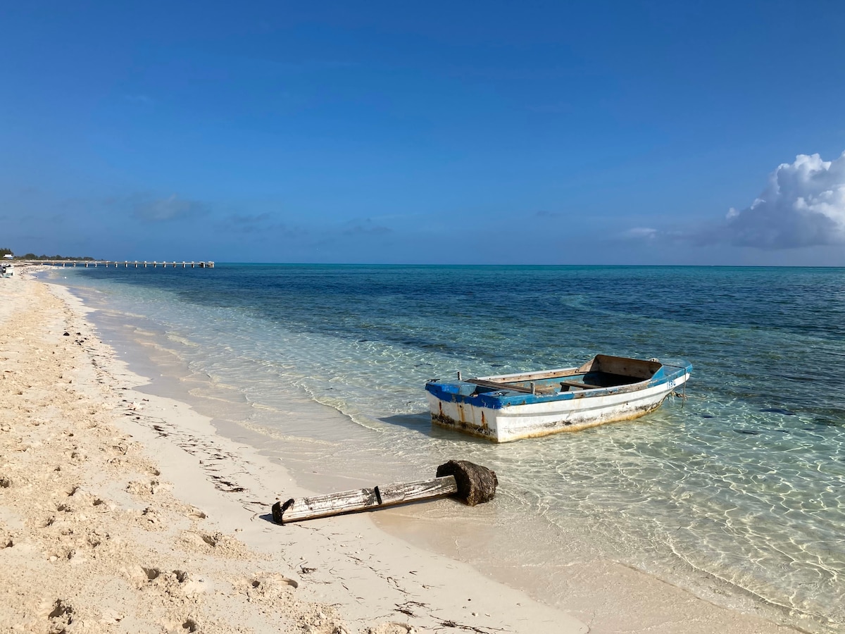 A small weathered boat is anchored gently in shallow water, positioned along a sandy beach. The clear turquoise sea stretches towards a blue sky with a few wispy clouds, creating a serene coastal scene.