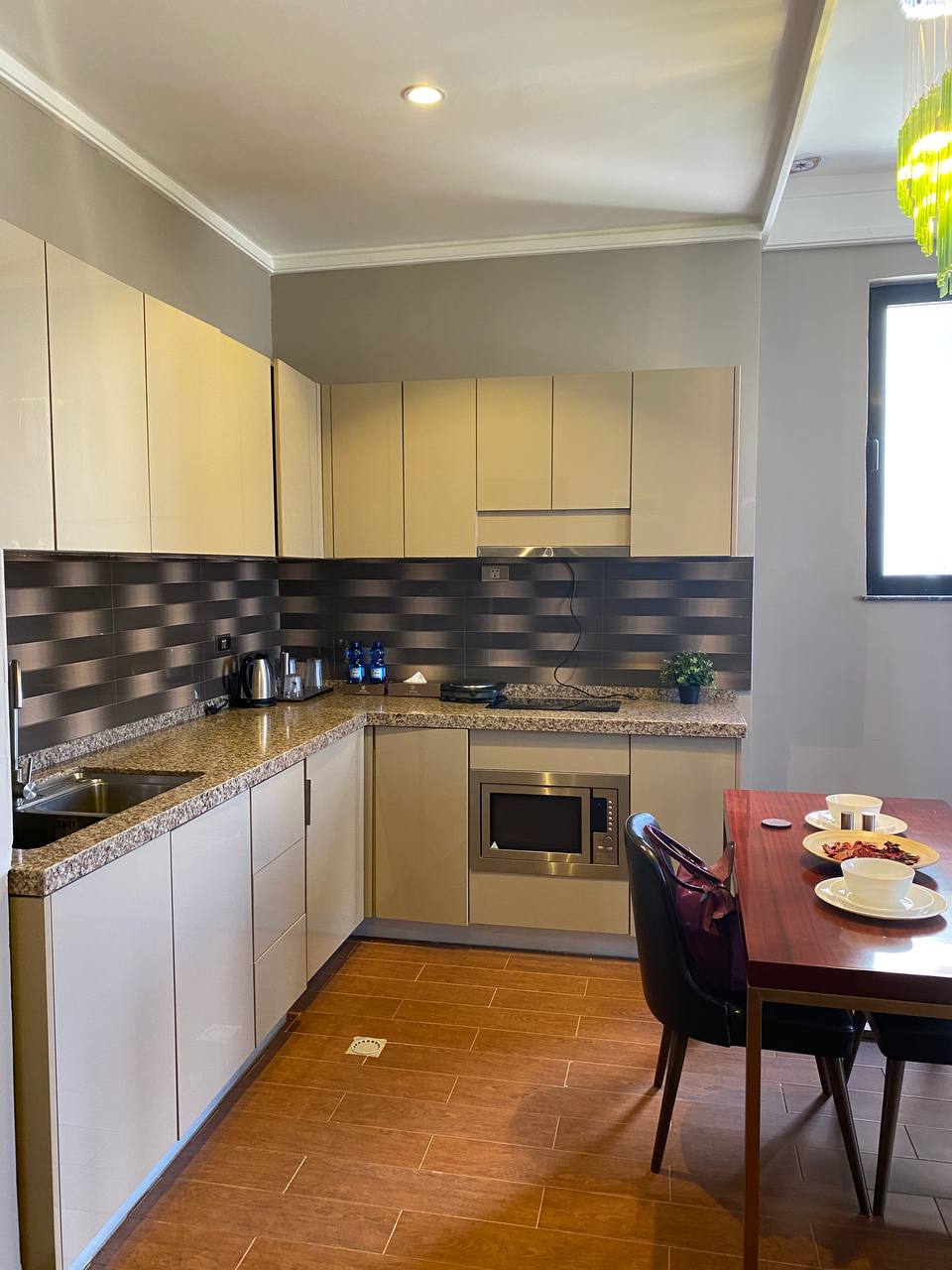 A modern kitchen area is equipped with sleek cabinetry and a granite countertop. Stainless steel appliances, including a microwave and electric kettle, are present. A dining table with two chairs is set with plates and fruit, while a light source enhances the warm tones of the flooring.