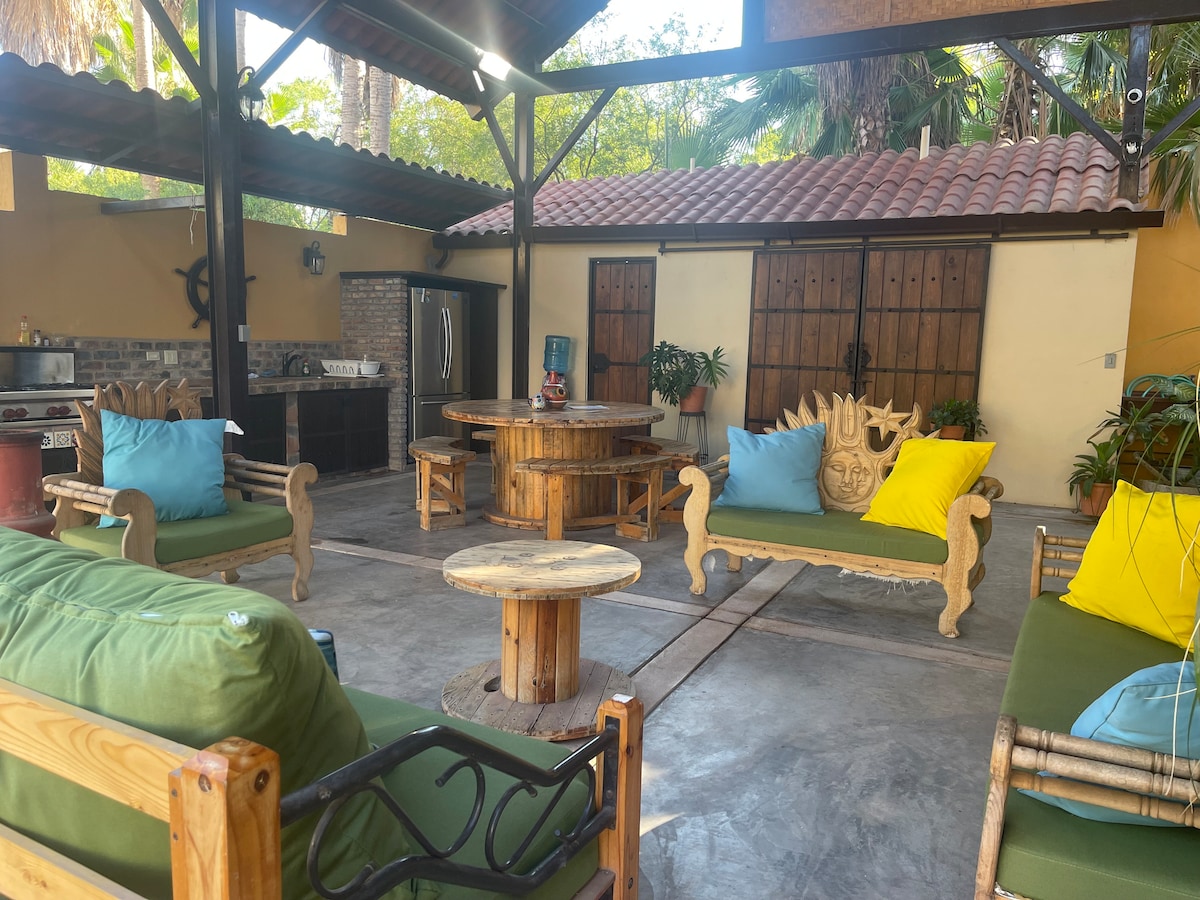 An outdoor lounge area features comfortable seating arranged around a circular wooden table. Vibrant yellow and green cushions adorn the furniture. A barbecue grill and kitchen area are visible in the background, surrounded by lush greenery and rustic wooden doors.