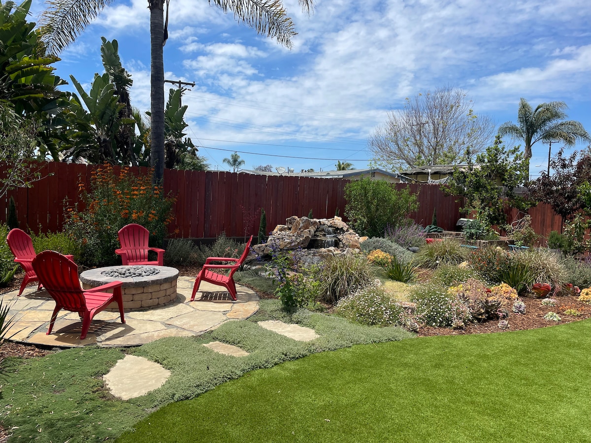 A serene garden area is revealed, featuring a stone fire pit surrounded by four red chairs. Lush greenery and colorful flowers are present throughout the space, with a gentle waterfall cascading in the background. Palm trees and a clear blue sky contribute to a peaceful outdoor setting.